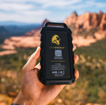 A hand holds a Lion Energy Staging Lion Cub portable charger outdoors, with red rock formations and blue sky blurred in the background.