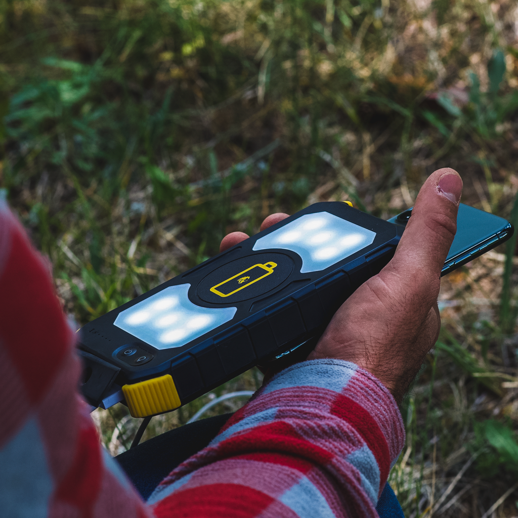 A person in a red plaid shirt holds a smartphone and the Lion Prowler portable power unit with LED lights and a battery icon by Lion Energy Staging, outdoors on grass.