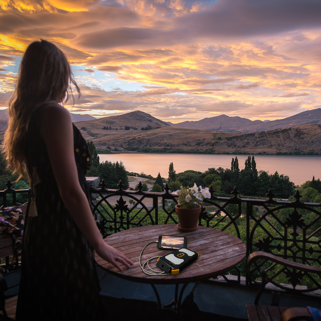 A woman stands on a balcony at sunset, overlooking a river and mountains. On the table in the foreground are a mobile device, charger, potted plant, and the Lion Energy Staging Lion Prowler portable power unit.