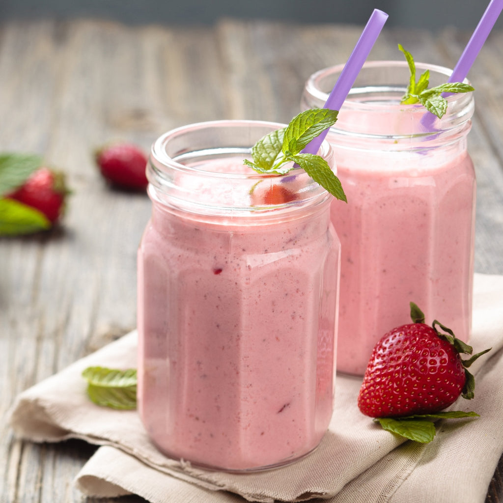 Two mason jars of pink strawberry smoothies, each with a purple straw and mint garnish, sit on a cloth napkin with fresh strawberries and Augason Farms Sliced Strawberries—a tasty vitamin C snack.